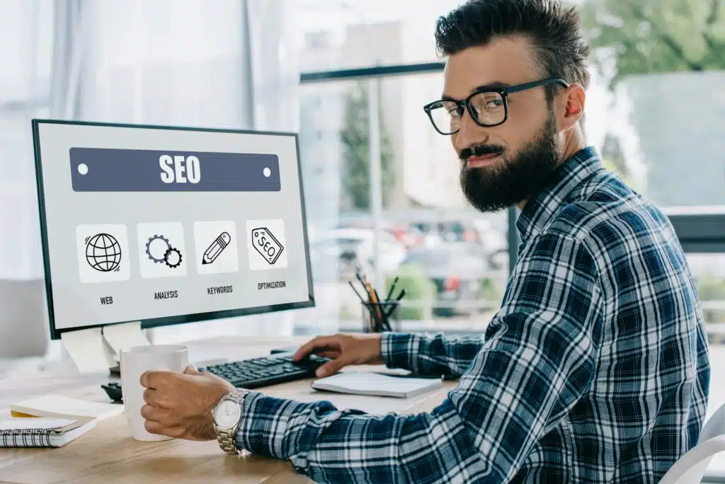 young successful seo manager sitting at workplace with computer and looking at camera